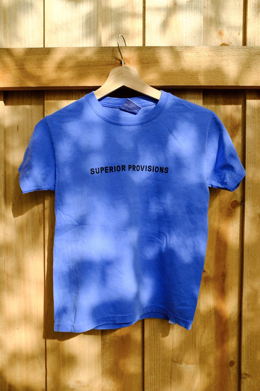 Kids blue shirt with "Superior Provisions" printed on the front, hanging from a wooden hanger in front of a wooden fence.