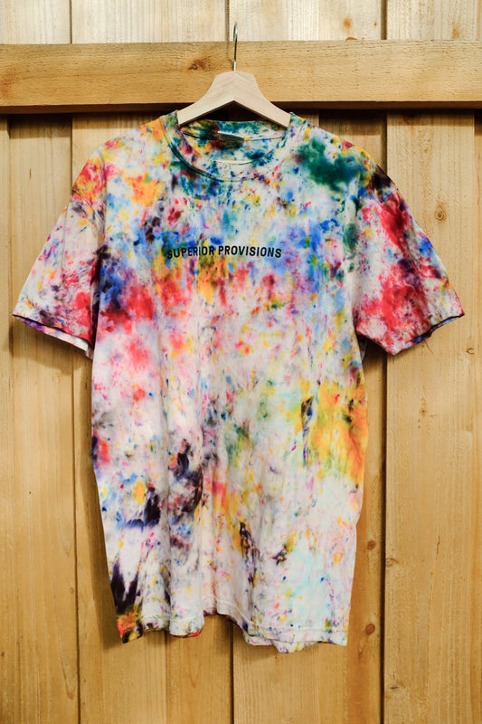 Tie-dye shirt with the "Superior Provisions" text printed in black on the front. The shirt is hanging on a wooden hanger in front of a wooden fence.