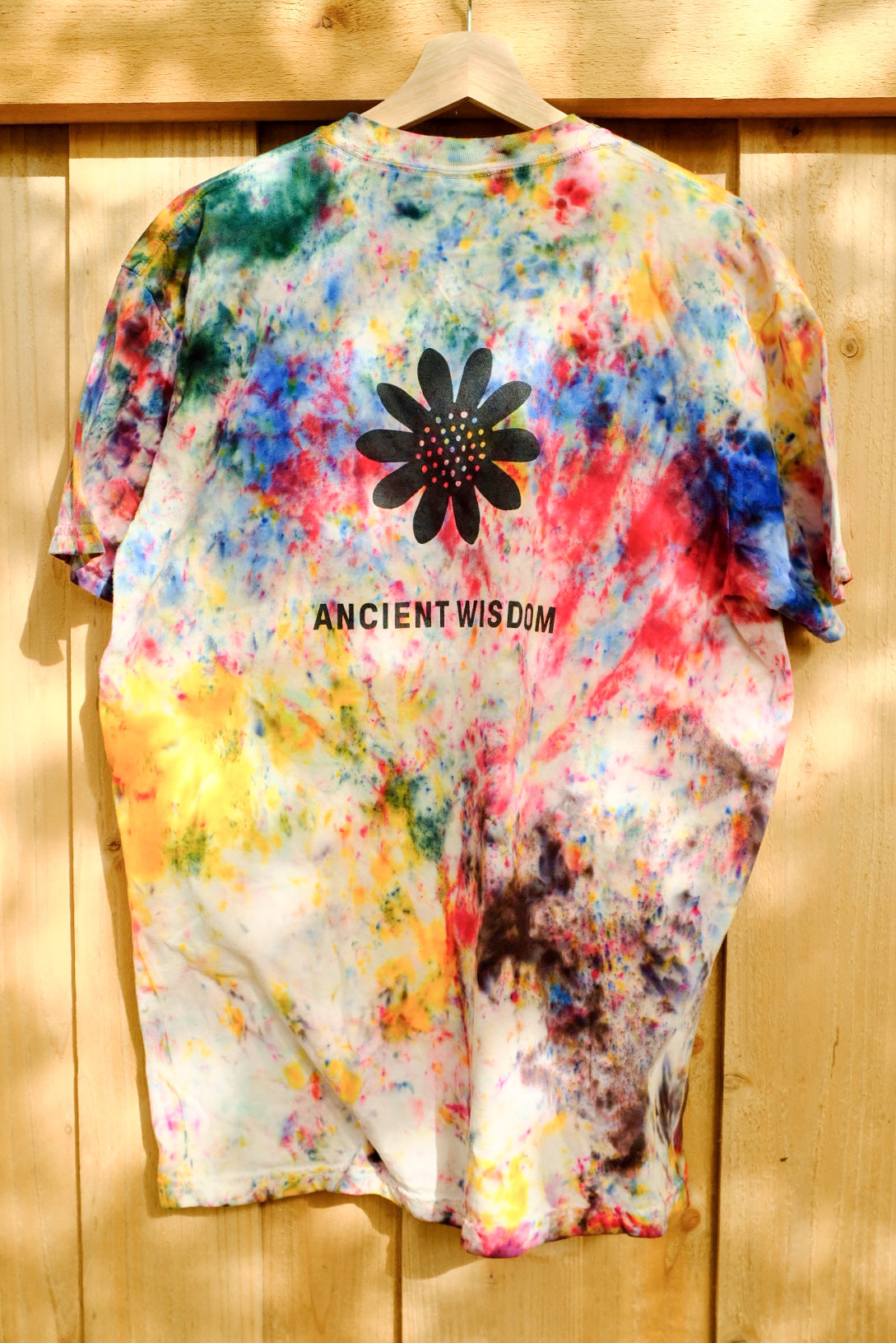 Tie-dye style shirt with "Ancient Wisdom" printed in black text underneath a black illustration of a sunflower. The shirt is hanging on a wooden hanger in front of a wooden fence.