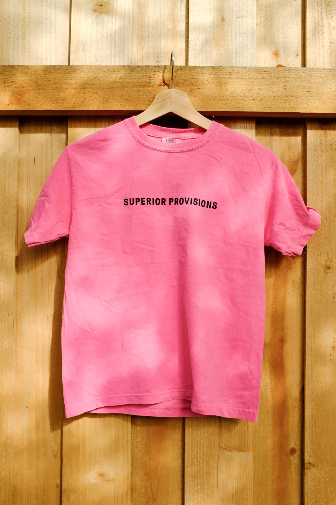 Front view of a pink t-shirt that has "Superior Provisions" printed in black. The shirt is hanging from a wooden hanger in front of a wooden fence.
