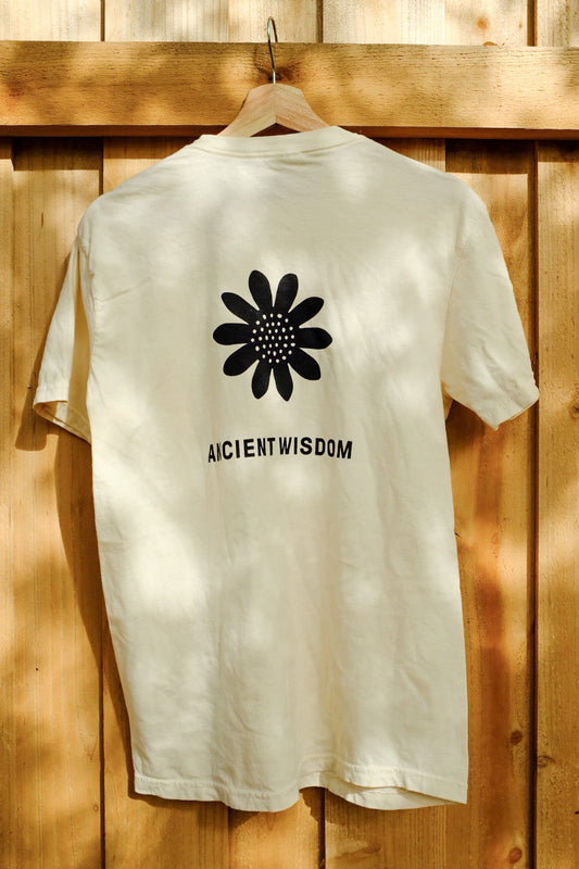 While shirt that says "Ancient Wisdom" in black text under a black illustration of a sunflower on a wooden hanger, in front of a wooden fence.