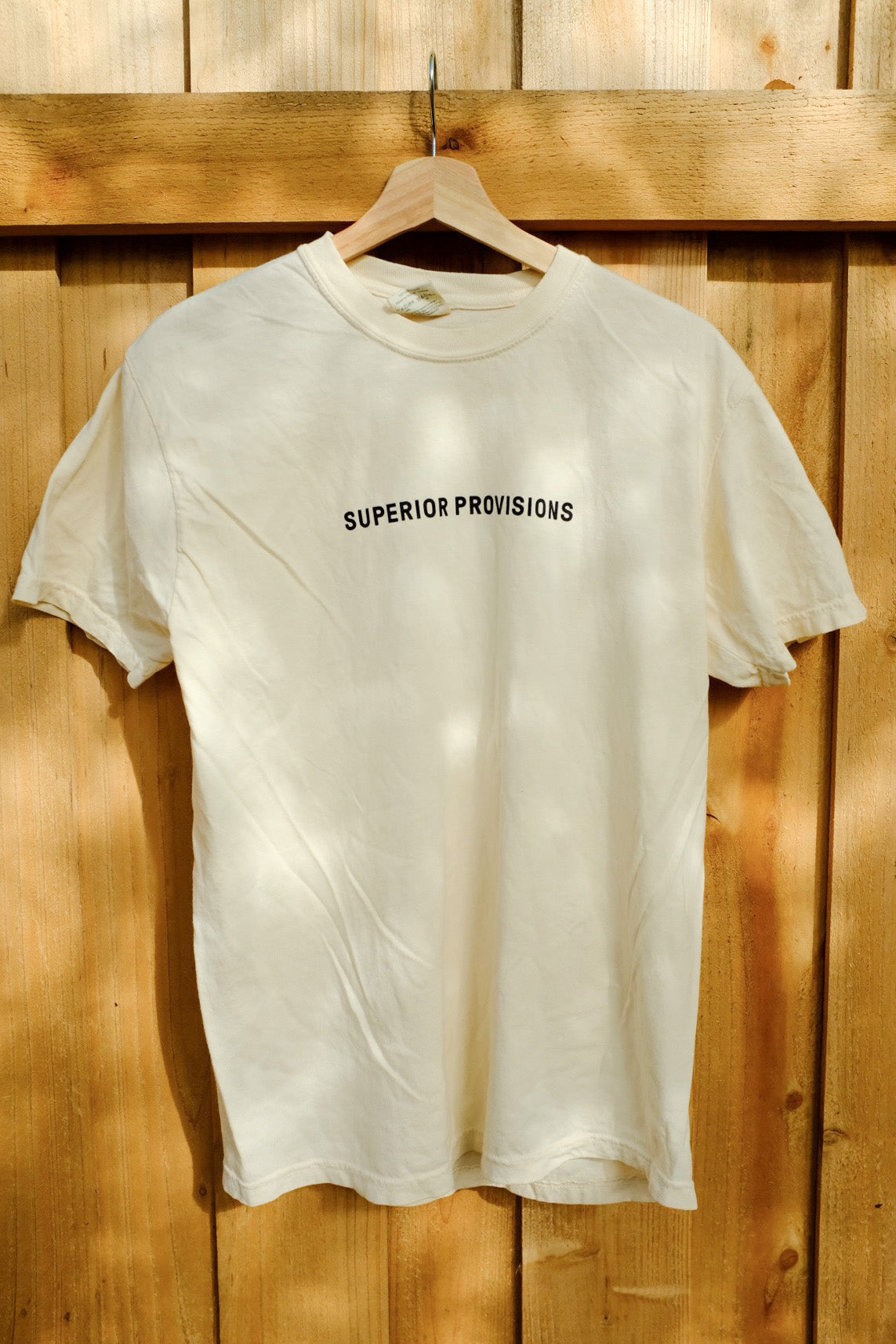 While shirt that says "Superior Provisions" in black text and is hanging on a wooden hanger, in front of a wooden fence.